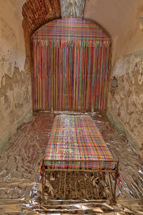 A blanket and curtain woven from multicolored shoelaces and mylar strips inside a crumbling cell