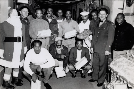 A group of prisoners and several guards stand in a cellblock near a Christmas tree and life size Santa decoration