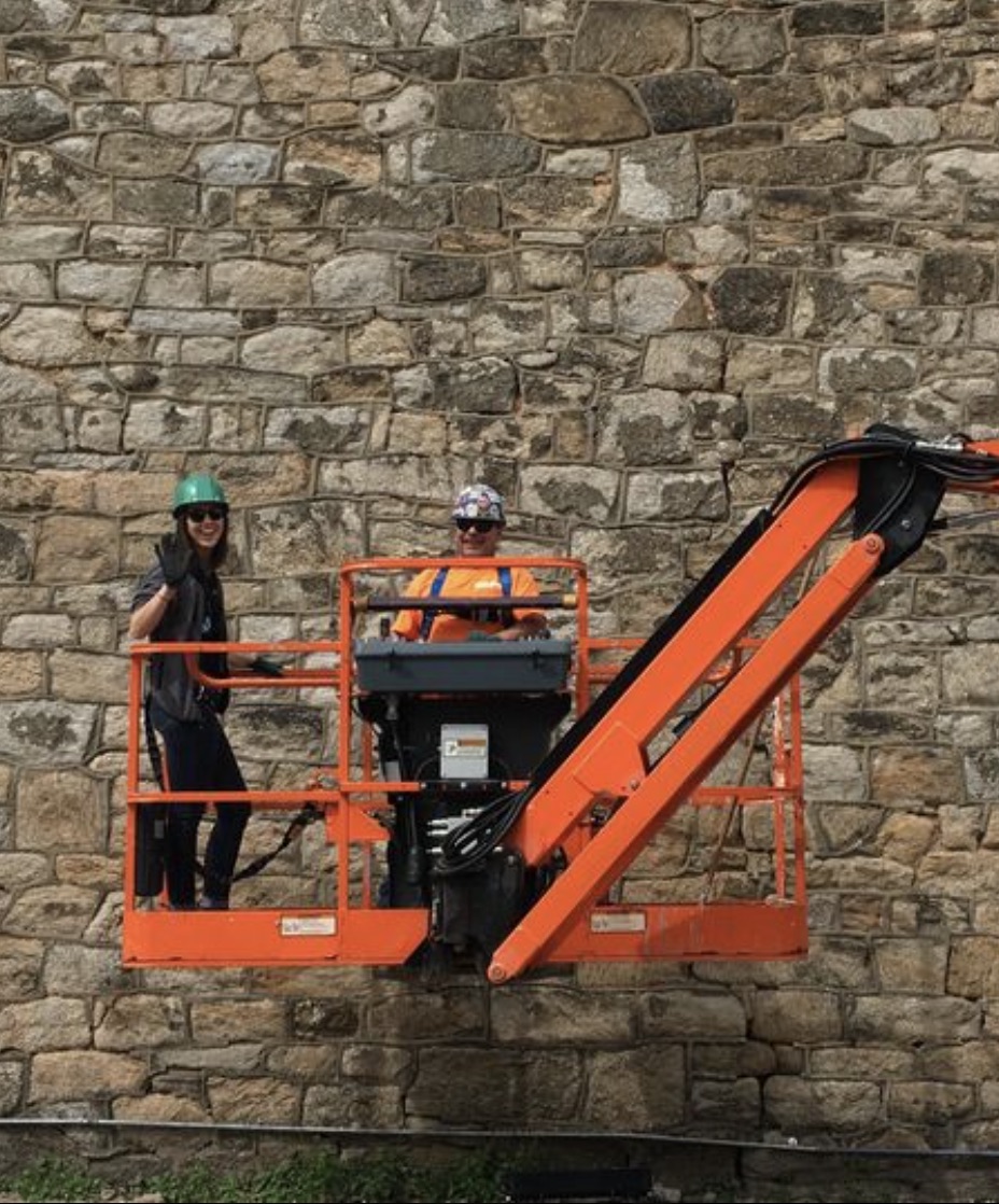 Two people in an orange basket lift, wearing hard hats, in front of Eastern State's historic stone wall 