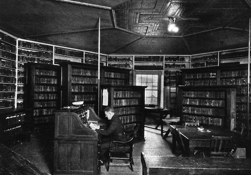 Black and white photo of man sitting at a large desk in a library, the room is shaped like an octagon and paneled in dark wood, books line the walls