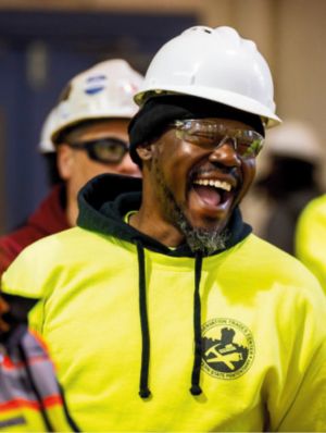 a man in a white hardhat, yellow sweatshirt, and safety goggles smiles 
