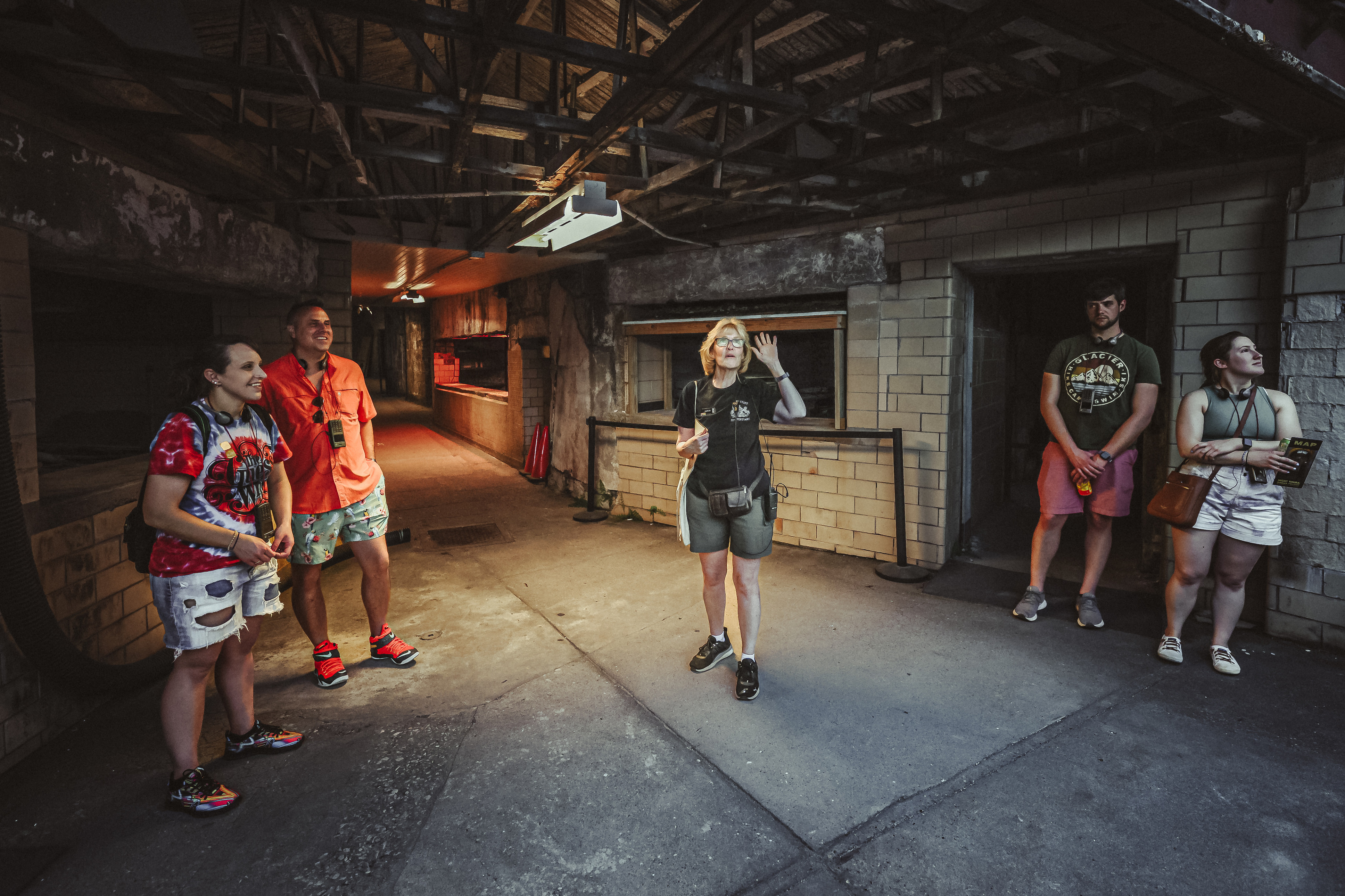 A photo of a mini tour at Eastern State Penitentiary. A tour guide is talking to a group of visitors who are looking at their surroundings; brick walls, a metal lattice ceiling, and red lightning in the background.