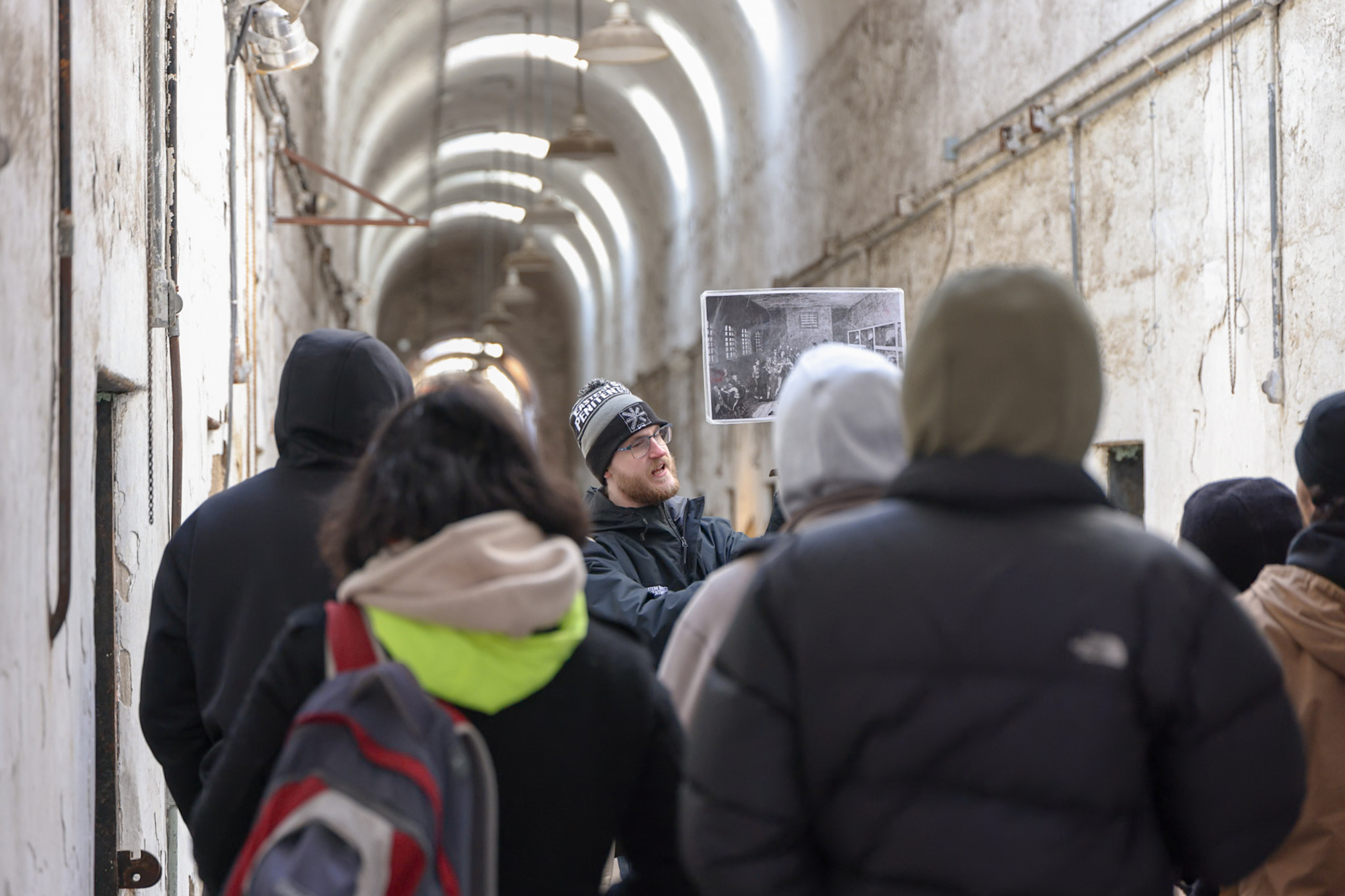 A photo of a group tour at Eastern State Penitentiary. A tour guide holds up a photo for a group of visitors. The background has peeling plaster and arched ceilings.
