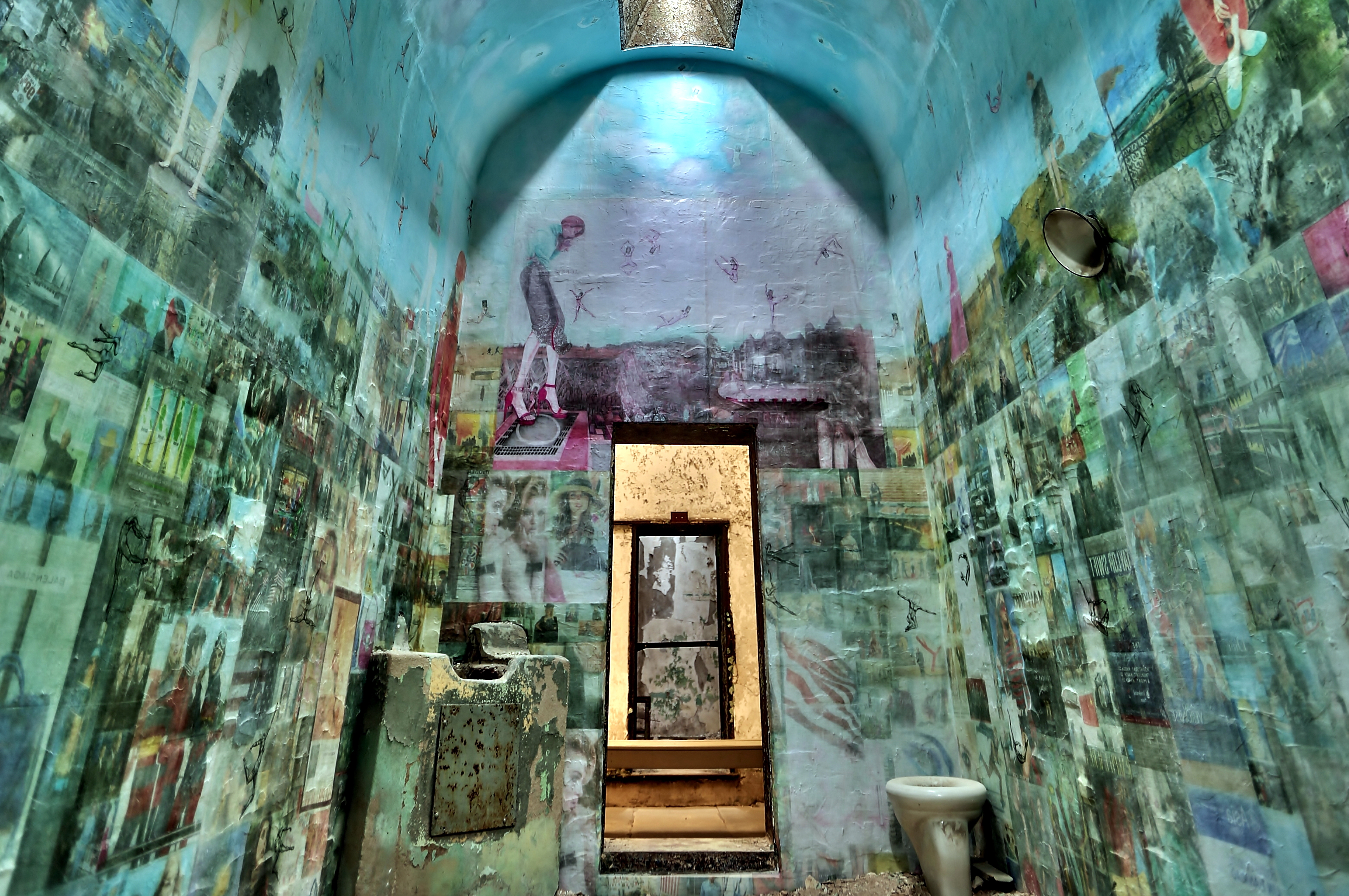 A photo of artist Jesse Krimes' exhibit Apokaluptein16389067:II at Eastern State Penitentiary. A cell is covered in photo collages across the walls and ceiling. The cell door is open showing the corridor outside it.