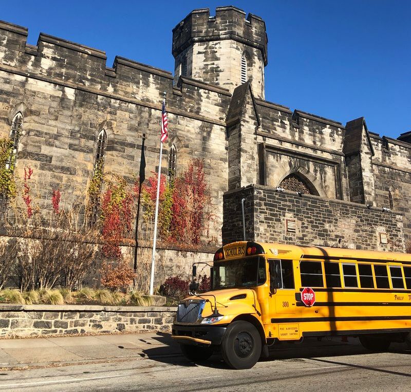 A photo of the facade of Eastern State Penitentiary. A school bus is parked on the street, the castle-like facade looking over it. There is also an American flag on a pole.