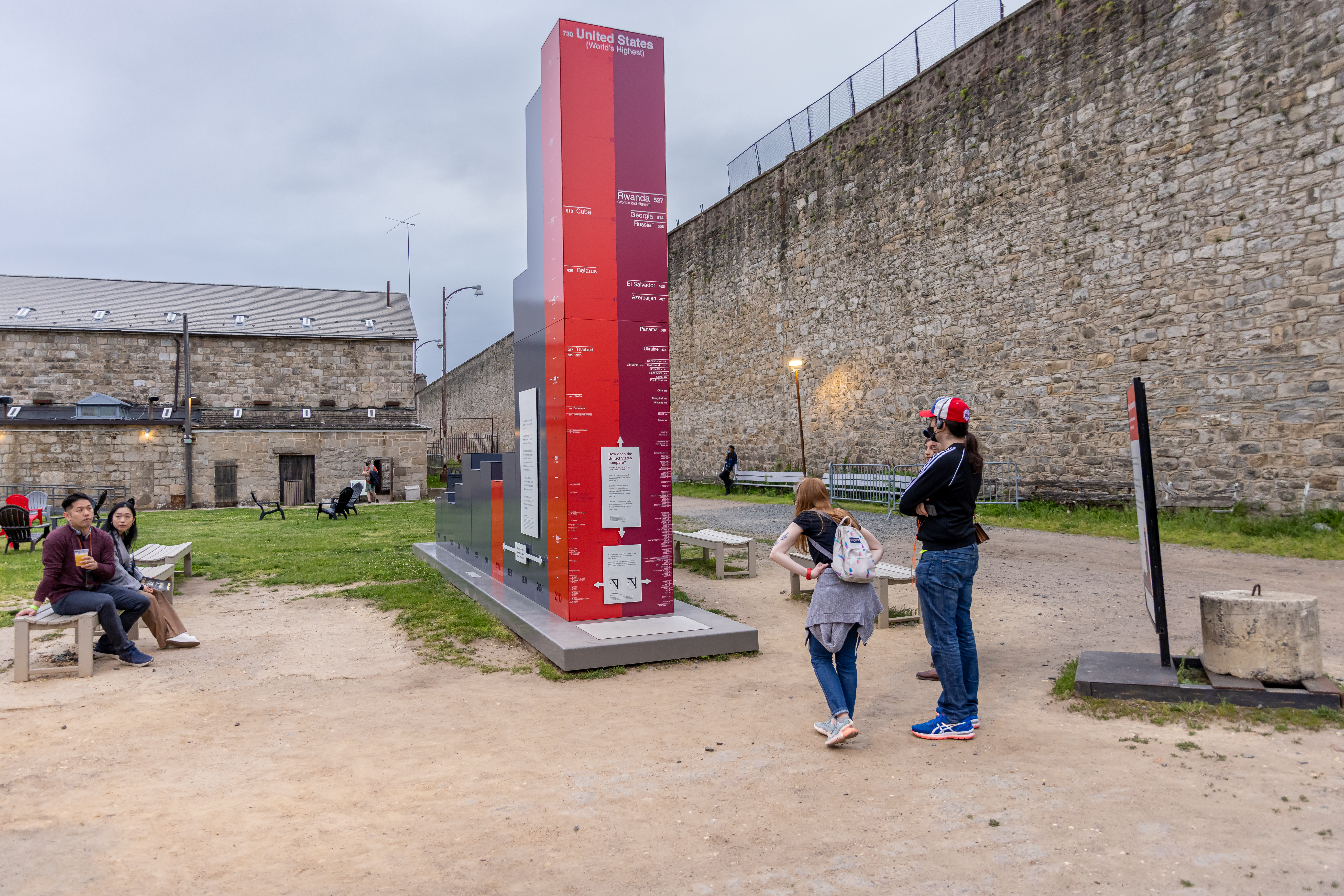 A photo of The Big Graph at Eastern State, a large outdoor bar graph sculpture, with people gathered around it observing and discussing the display. The tallest bar is red and at the top shows the United States as having the highest incarceration rate in the world.