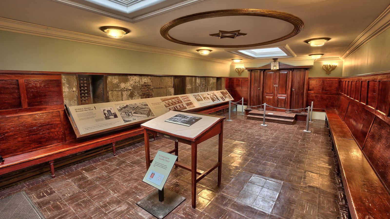 A photo of the synagogue at Eastern State Penitentiary. There is wooden paneling on the walls and benches lining the sides. A wooden table is visible, and at the far end is a wooden cabinet, the Torah Ark. The ceiling features a circular design with a Star of David in the center.
