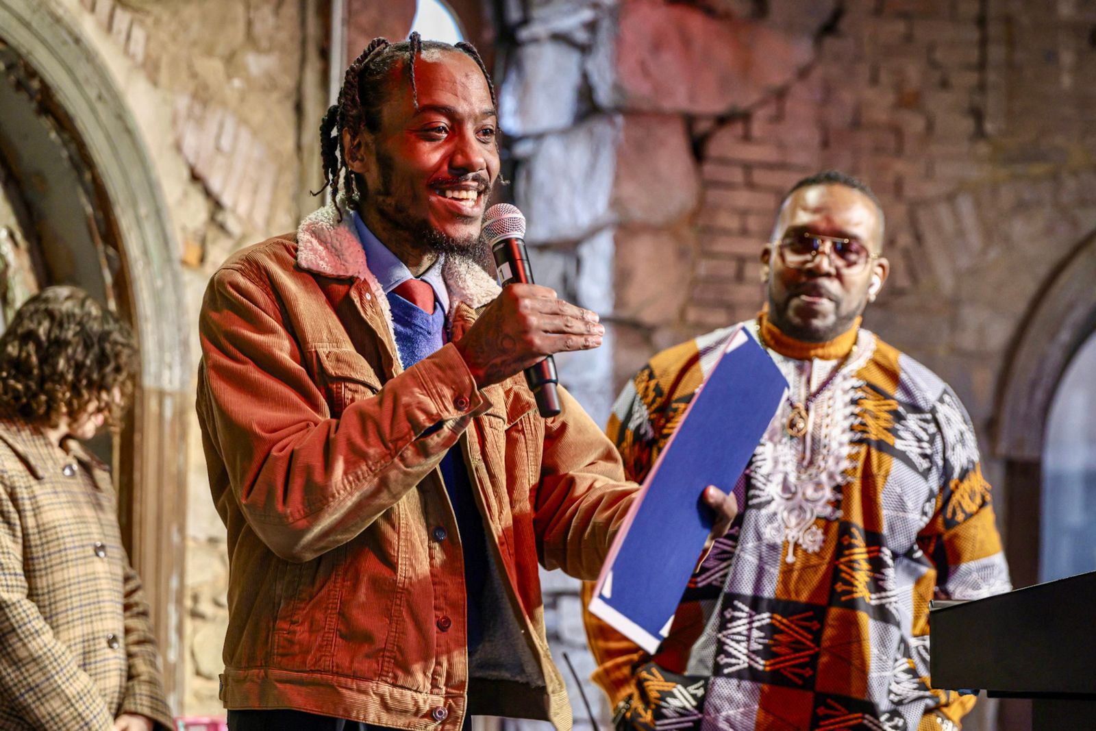 A photo of a Preservation Trades Center graduate talking at Eastern State Penitentiary. The speaker holds a microphone. He is set against aged stone and brick walls.